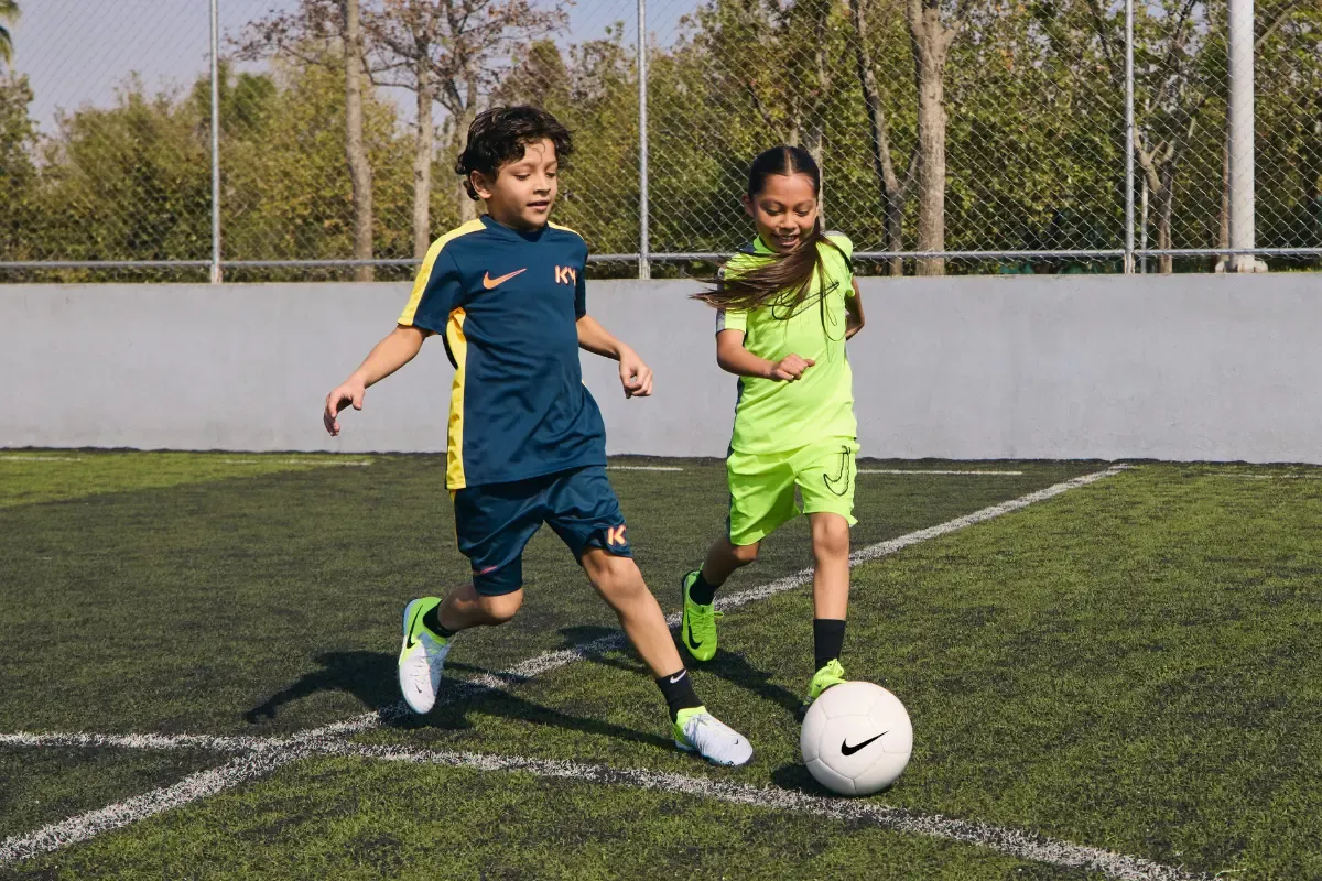 Duas crianças jogando futebol em campo usando uniforme e chuteiras.