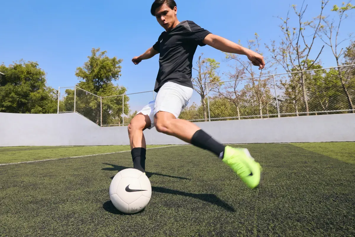 Jogador chutando bola de futebol em campo usando chuteiras Nike.