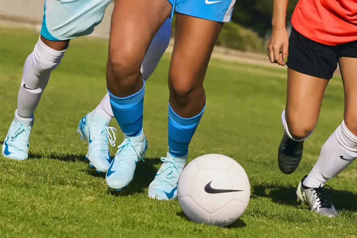 Jogadores de futebol em ação durante uma partida, com foco nas pernas e chuteiras, disputando a bola Nike em um campo de grama.