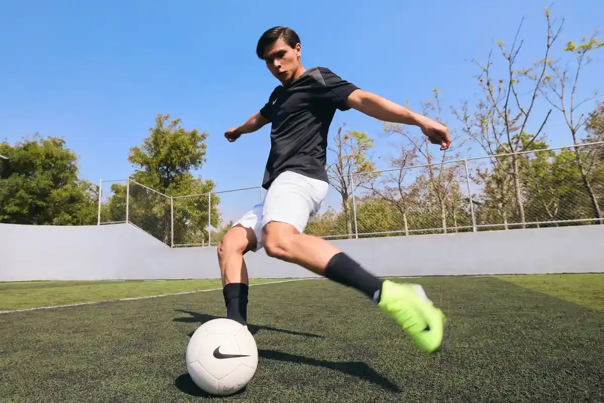 Jovem jogador de futebol praticando drible em campo ao ar livre em dia ensolarado, com céu azul e árvores ao fundo.