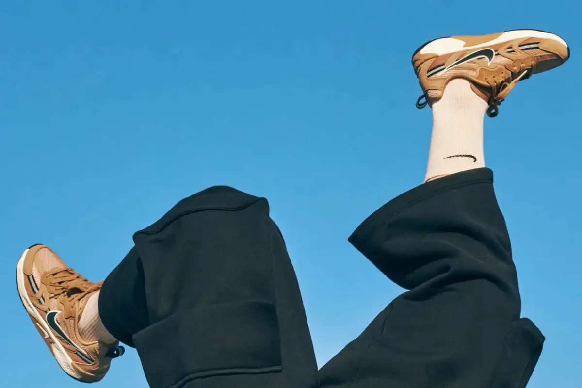 Pessoa brincando e levantando as pernas ao ar com tênis esportivos, usando calça preta, contra um céu azul claro.