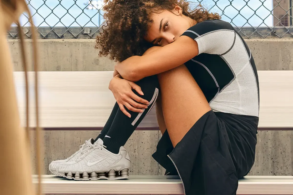 “Mulher com roupa esportiva sentada em cerca de quadra usando tênis Nike.”