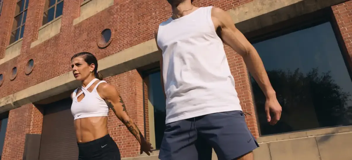 Duas pessoas fazendo exercícios ao ar livre na área urbana, destacando o fitness e a saúde, com uma mulher com abdominal definido e um homem usando roupas de ginástica.