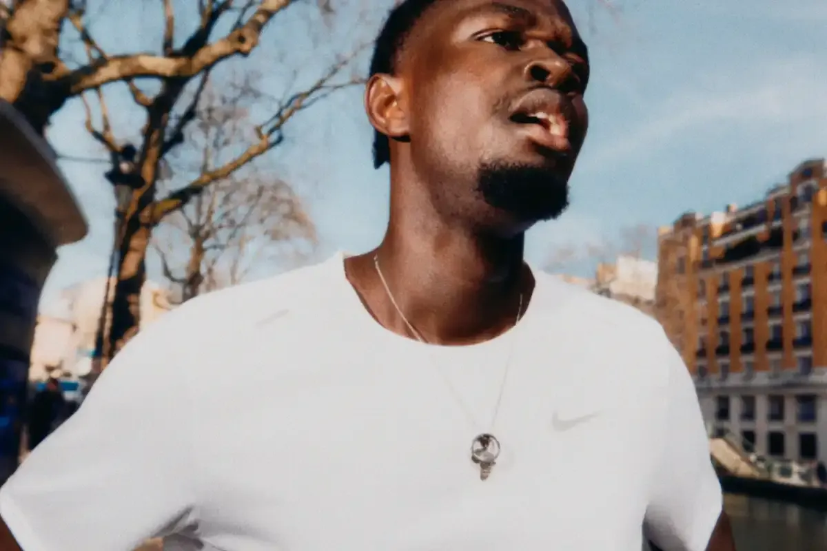 Homem com cabelo curto e barba, usando uma camiseta branca e uma corrente com pingente, ao ar livre em uma cidade, possivelmente durante o dia.