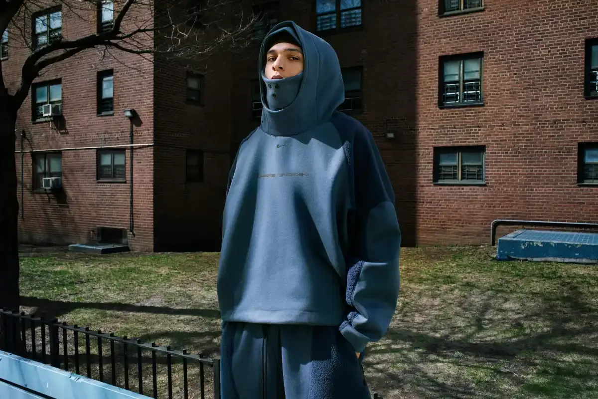 Jovem usando moletom oversized com capuz na rua de um bairro residencial, com prédio de tijolos ao fundo e sombra de árvores no chão.