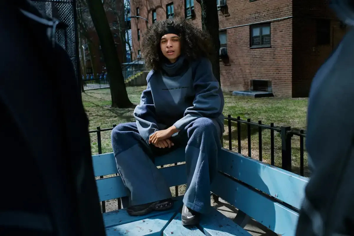 Jovem de cabelos cacheados e estilo casual sentado em banco de parque urbano, com prédios ao fundo, refletindo uma cena de rua moderna.