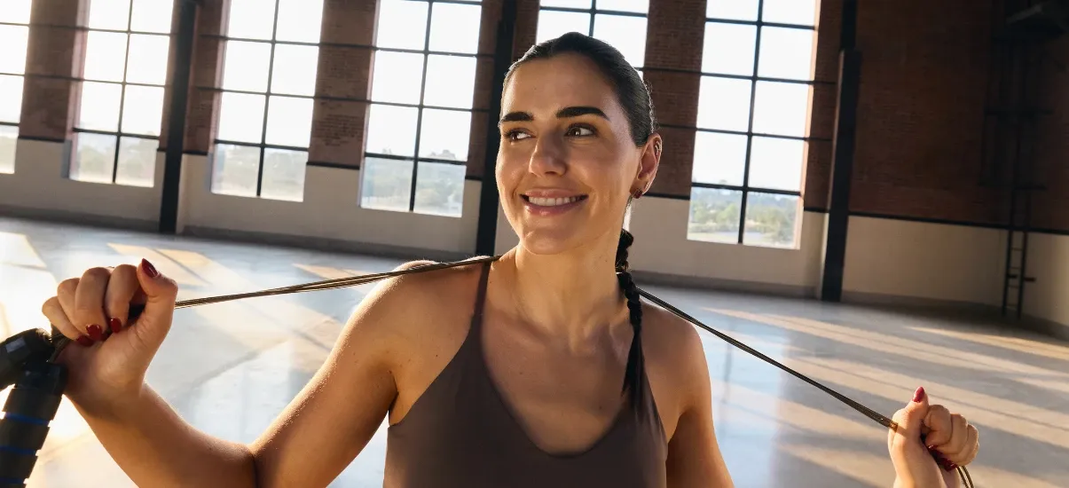 Mulher fazendo exercício de alongamento com faixa de resistência em ambiente interno bem iluminado e com janelas grandes ao fundo, promovendo saúde e bem-estar.