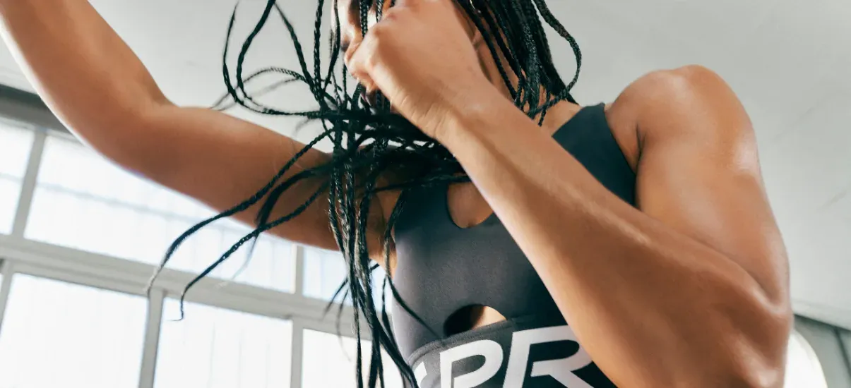 Mulher em movimento durante um treino, usando top esportivo cinza com recorte frontal e faixa com logo na barra.